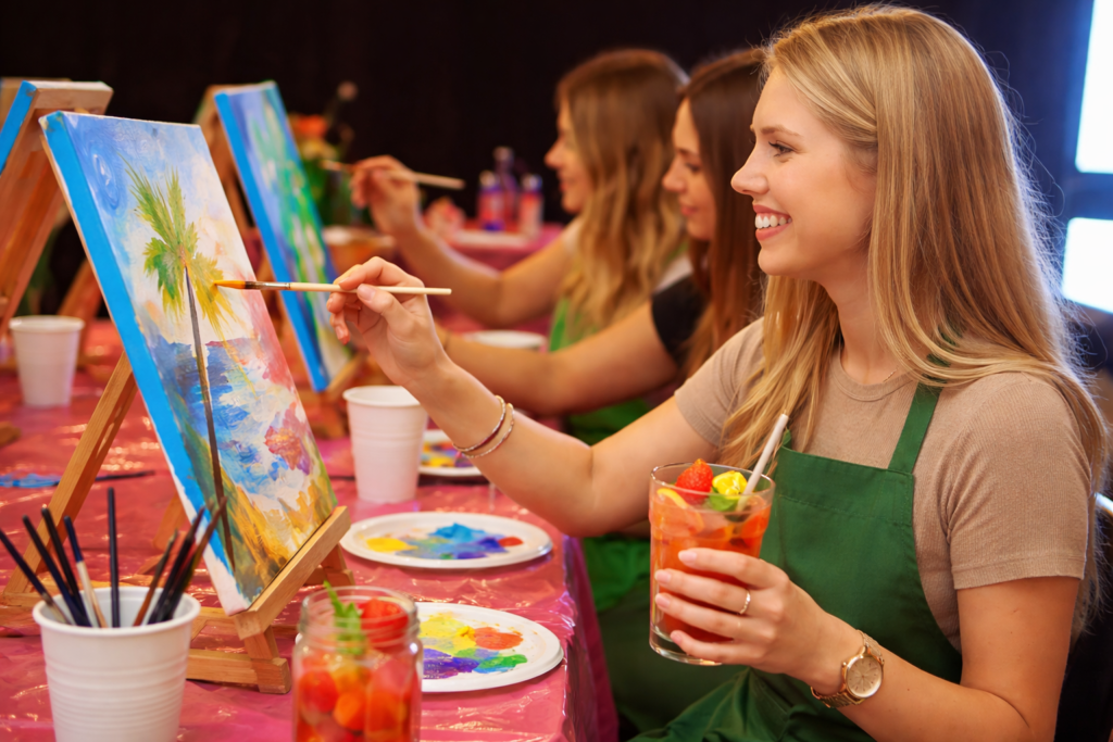 Three women painting on canvas while holding mocktails.